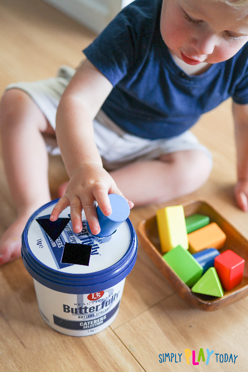 Child slides a round block into the circle cutout in the lid of a plastic container. There is also a square and a triangle cutout in the lid for the other blocks